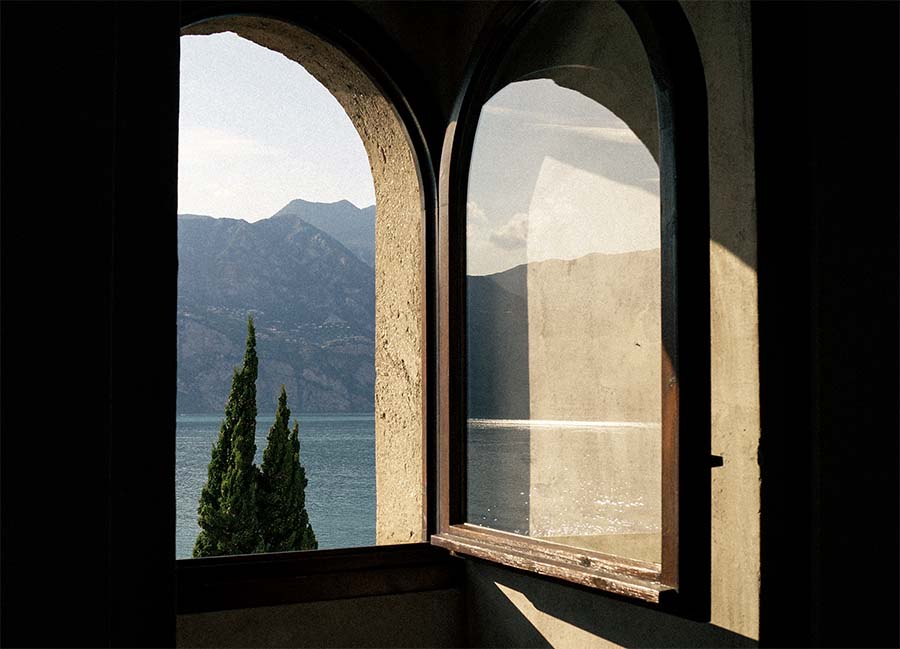 Cover image for Between the Shadows album, an open window in a dark room lets sunlight in from outside, lake and mountains in the distance
