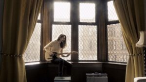 Jonni Slater sitting in a sunny bay window playing a guitar. 