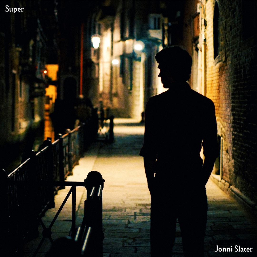 Cover image of the single Super. A silhouetted figure of Jonni Slater standing in a darkened street in Venice, with streetlights creating pools of light along the cobbled walkway that recedes into the distance.