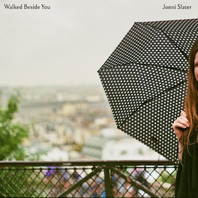 Cover image for Walked Beside You single. A figure stands just out of shot with only half of their umbrella visible, a cityscape out of focus in the background, with the camera showing the empty space beside the figure.
