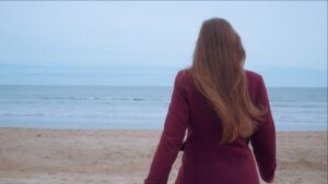 Frame from the video of Walked Beside You - a female figure in a purple coat walks away from the camera along a sandy beach towards the blue sea and sky.